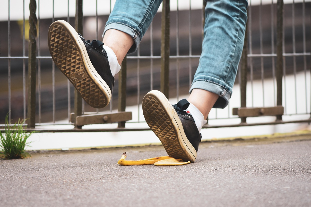 Close-up of a person wearing rolled-up jeans, white socks, and black sneakers stepping on a banana peel on the pavement, with both feet mid-air as if slipping. A metal fence is visible in the background.