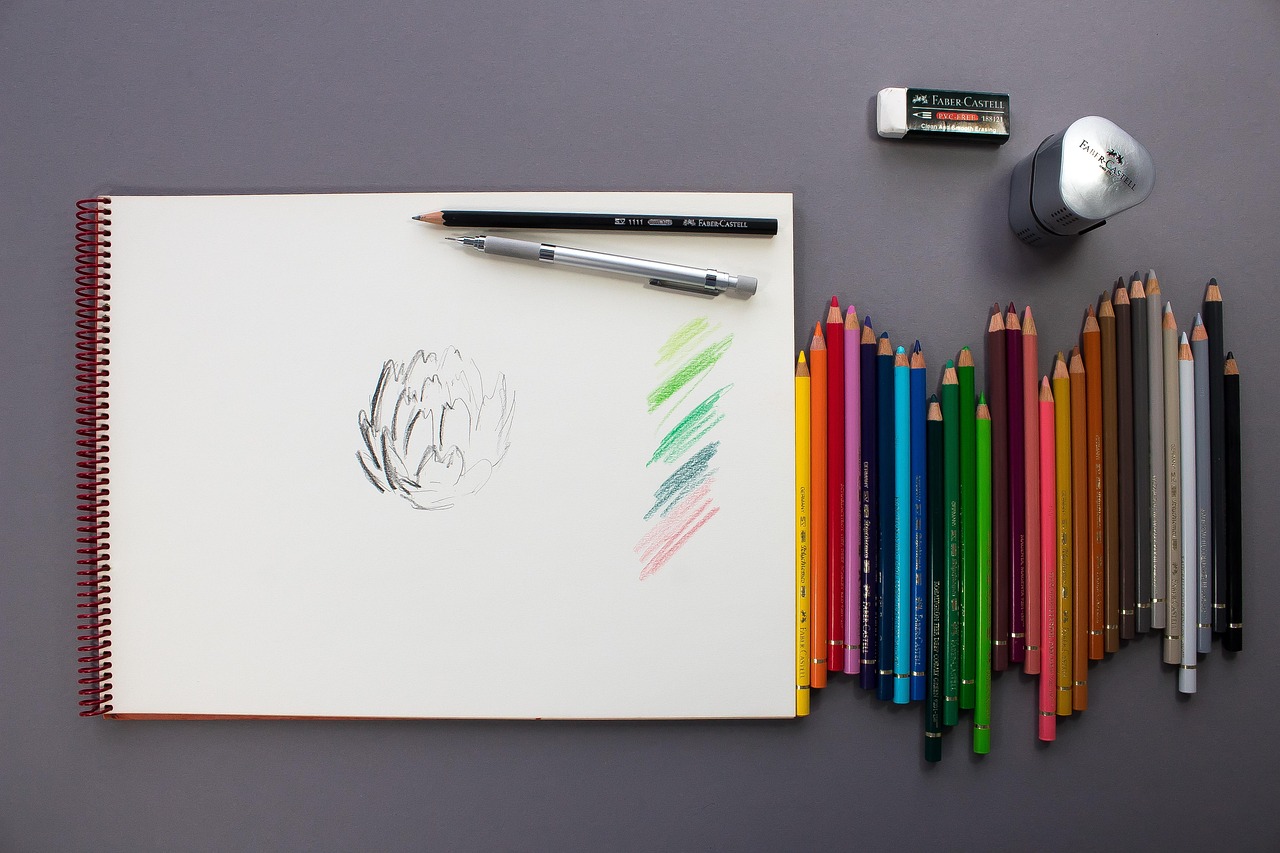 Sketchpad with a light sketch of an artichoke and several colored pencil lines. Beside the pad are a number of colored pencils, a pencil sharpener, and an eraser