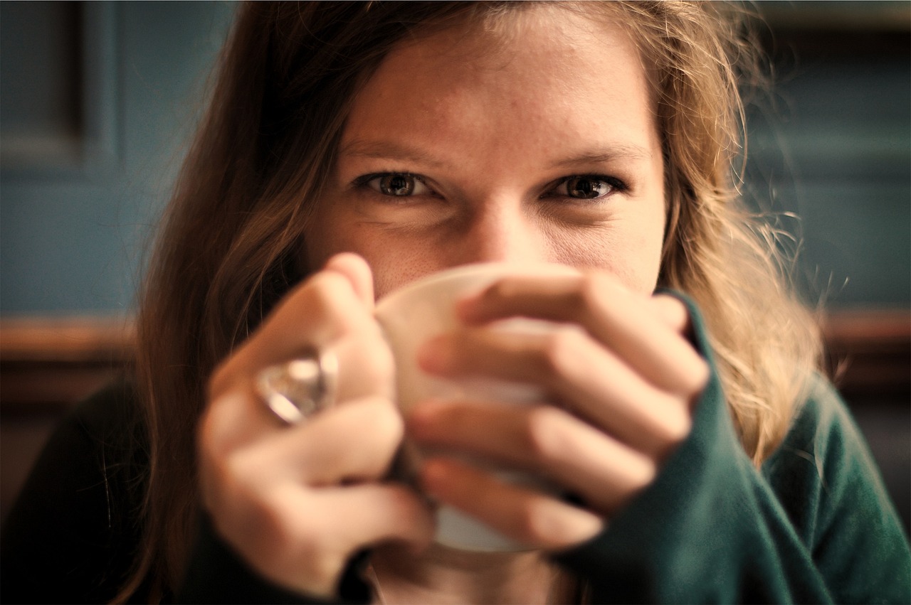 Close-up of a smiling woman with long, light brown hair holding a white mug with both hands, wearing a green sweater, looking directly into the camera.